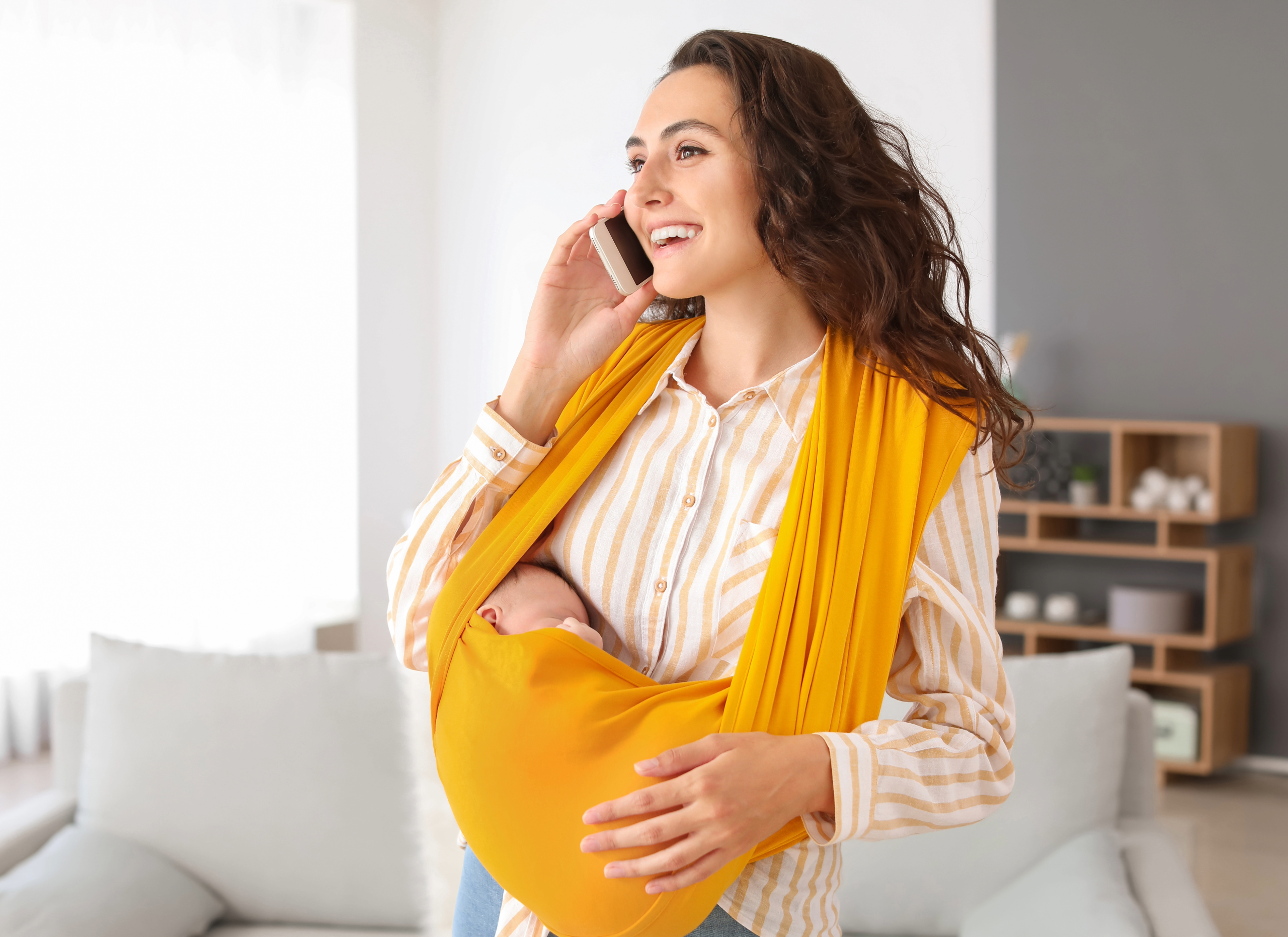 Mother speaking on the phone with a yellow baby carrier in the livingroom